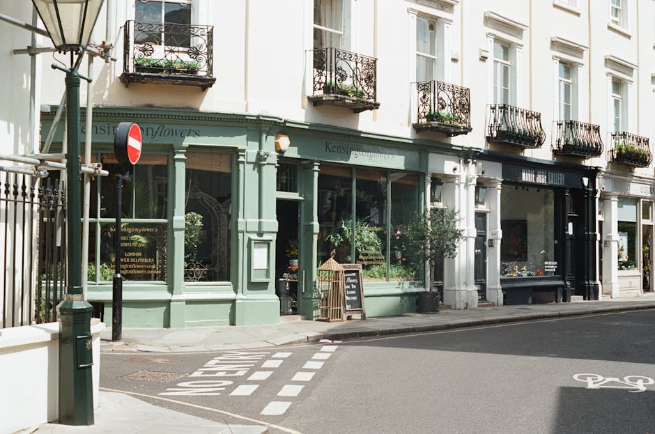 The image depicts the exterior of a flower shop located on a corner in Maida Vale, Elgin Avenue. The shop features a charming light green facade with large glass windows displaying various floral arra