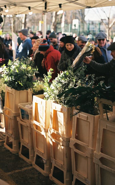 A busy outdoor flower stall featuring fresh floral bouquets arranged in wooden crates under a canopy, with a diverse crowd of people browsing and selecting flowers. The arrangement includes vibrant gr