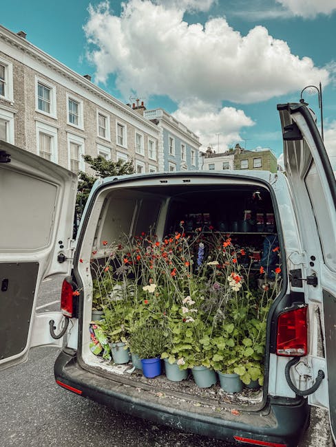 A white delivery van with its rear doors open, showcasing a variety of freshly cut flowers in pots and small containers, including red poppies, white daisies, and green foliage, arranged on the floor