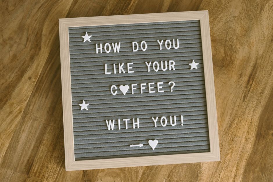 A square felt letter board with a light wooden frame displaying white plastic letters spelling out 'How do you like your coffee? with you'. The board is placed on a wooden surface with a warm honey to