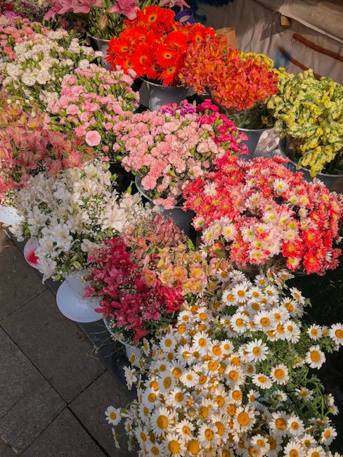 A vibrant display of fresh flowers arranged in various containers at a flower market, featuring a diverse selection of blooms including white and pink chrysanthemums, daisies with yellow centers, brig