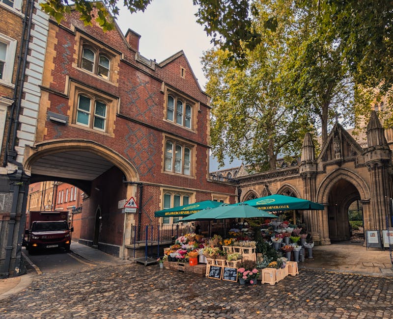 A street scene featuring a flower stall set up under a large green umbrella on cobblestone pavement, displaying an assortment of fresh flowers in various colors including pinks, oranges, and whites, a