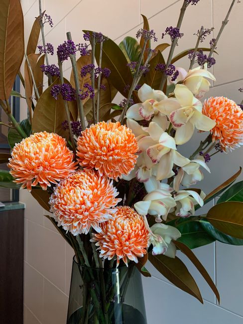 A tall, elegant floral arrangement featuring a combination of large, round, orange and white carnations with densely petaled blooms, complemented by cascading white orchids with pink accents, all set 