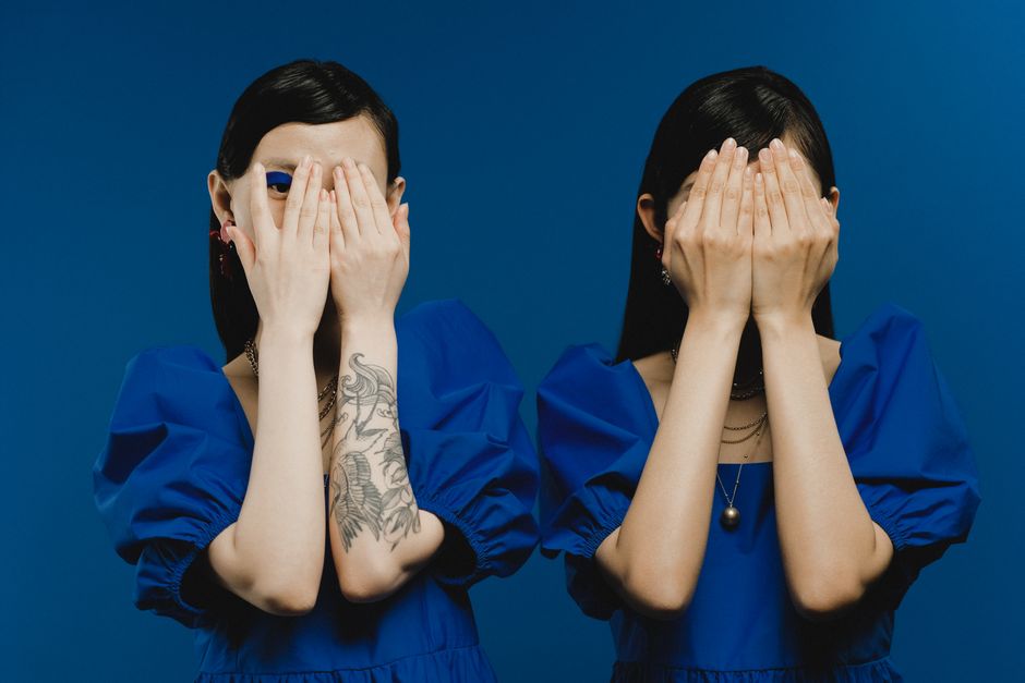 Two women with dark hair and pale skin wearing matching royal blue dresses with puffed sleeves, standing against a solid blue background. Both women are covering their faces with their hands, each hol
