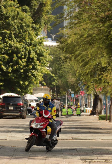 There is no image of flowers, floral arrangements, or related materials in the provided photograph. The scene depicts a city street with trees, parked cars, and a delivery person riding a red scooter