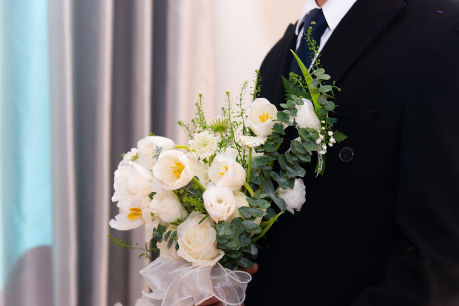 Close-up of a man in a formal black suit holding a floral bouquet featuring white tulips, white roses, and delicate white lisianthus, accented with eucalyptus leaves and small white filler flowers. Th