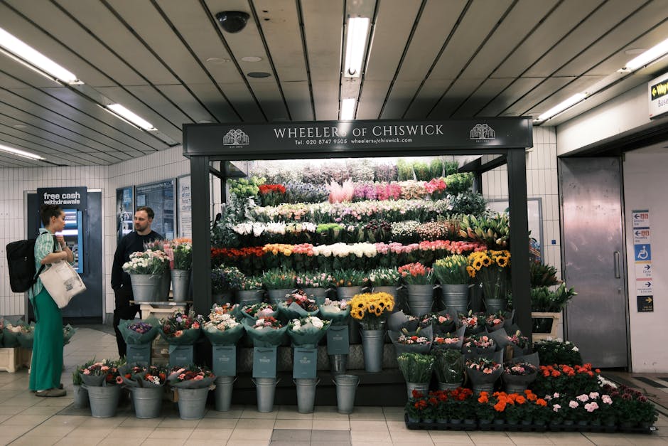 A vibrant floral display at Wheeler of Chiswick in an underground station, featuring an array of freshly cut flowers arranged in grey buckets and wrapped bouquets. The arrangement includes bright yell
