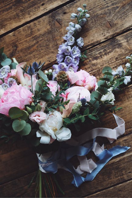 A floral bouquet arranged with soft pink peonies, white and pale purple stock flowers, and sprigs of eucalyptus with rounded green leaves, all tied together with satin ribbons in shades of white and p