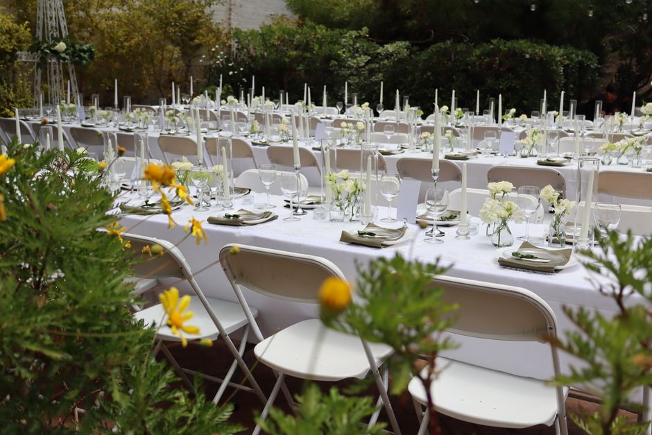 A long outdoor banquet table set for a wedding with elegant floral arrangements featuring white and cream flowers in small glass vases, complemented by tall white taper candles in silver candlesticks.