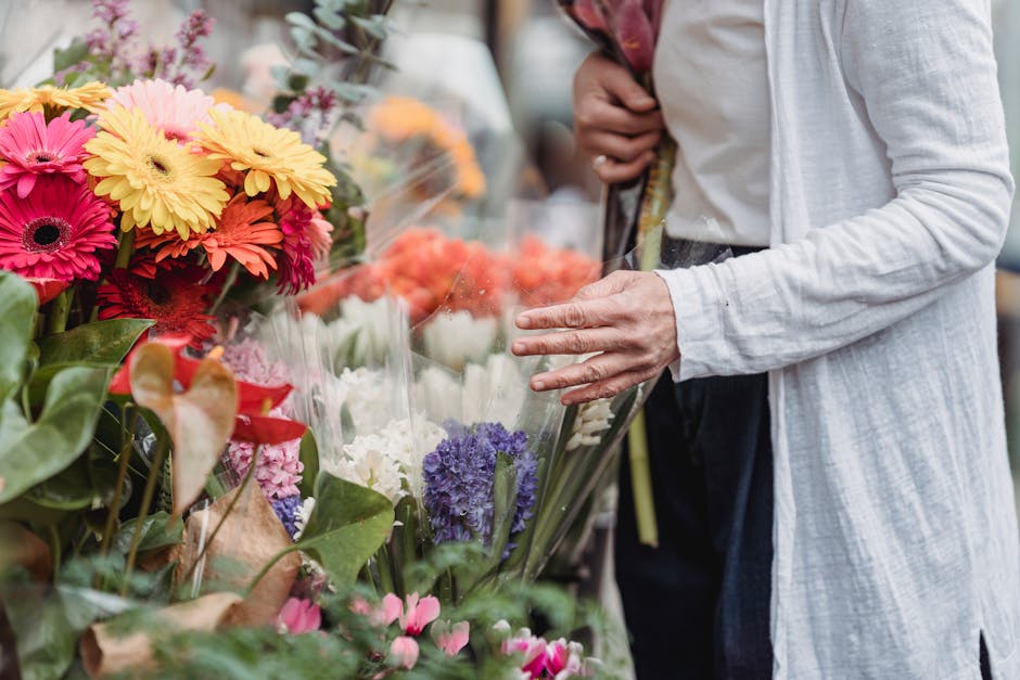 A person wearing a light grey, long-sleeved cardigan is selecting fresh flowers from a floral display at a florist. The bouquet includes vibrant pink, yellow, and orange gerbera daisies, purple hyacin