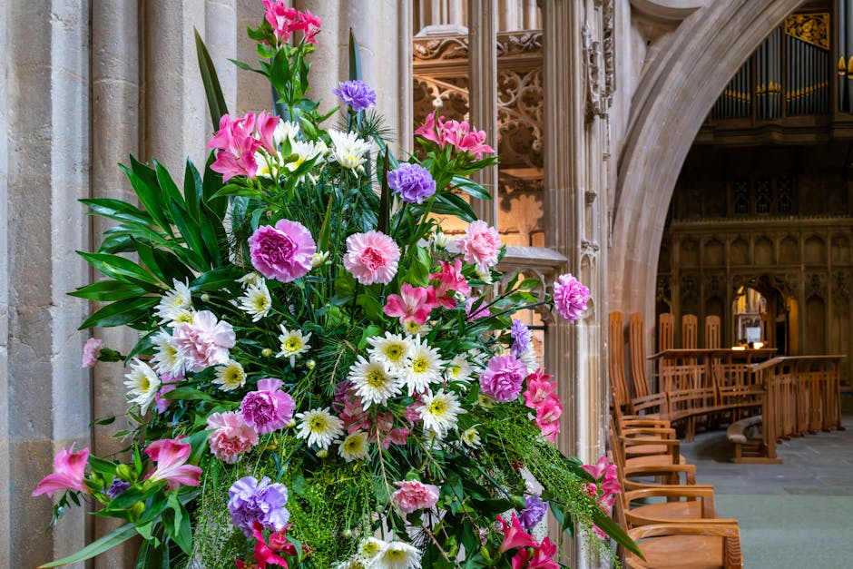 A large floral arrangement featuring pink lilies, purple and pink carnations, white daisies, and greenery placed inside a historic building with stone walls and wooden pews. The bouquet is lush and vi